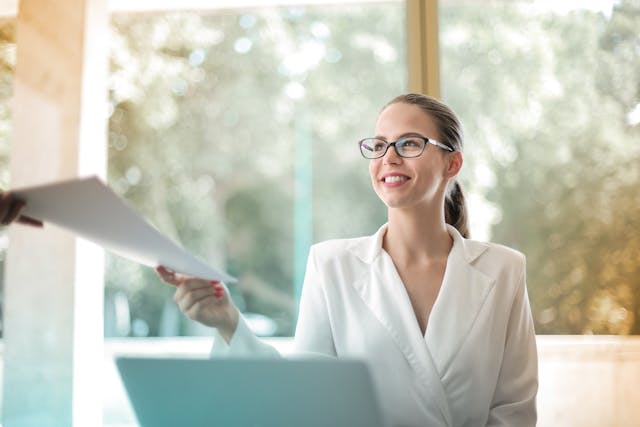 a woman in a white blouse and glasses in front of a laptop receiving a stack of papers from someone else